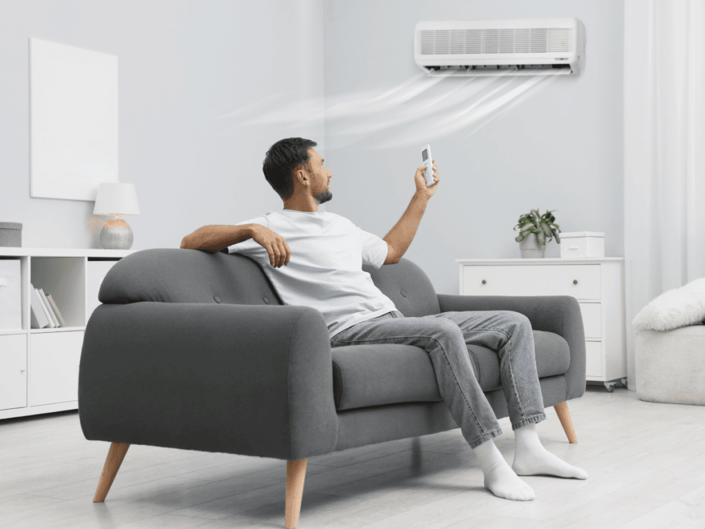 A man sits on a gray sofa using a remote to control a wall-mounted air conditioner, enjoying cool comfort in his bright, modern living room during the seasonal summer heat.