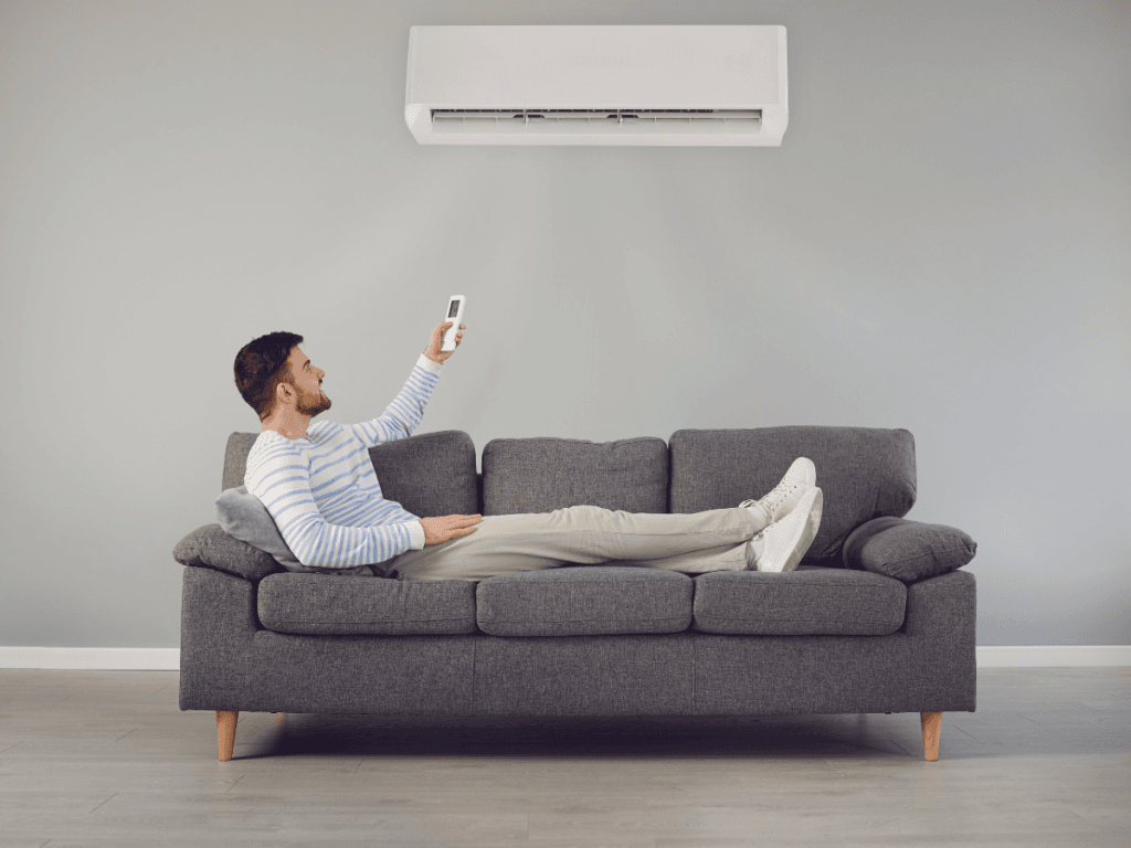 A man lying on a gray sofa uses a remote control to operate an air conditioner mounted on a plain gray wall, appearing frustrated as the AC is not cooling the room properly.