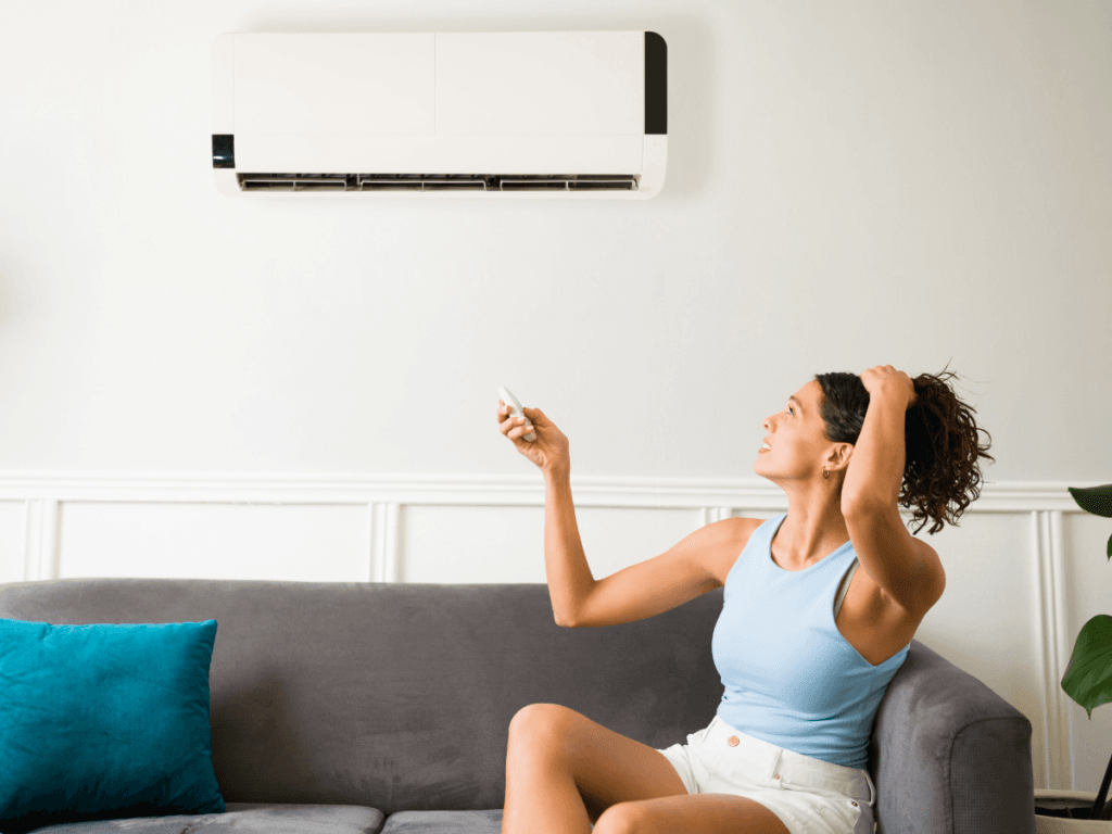 Woman sitting on a gray sofa uses a remote to adjust a wall-mounted air conditioner while looking up at the unit, appearing concerned about the AC not cooling.