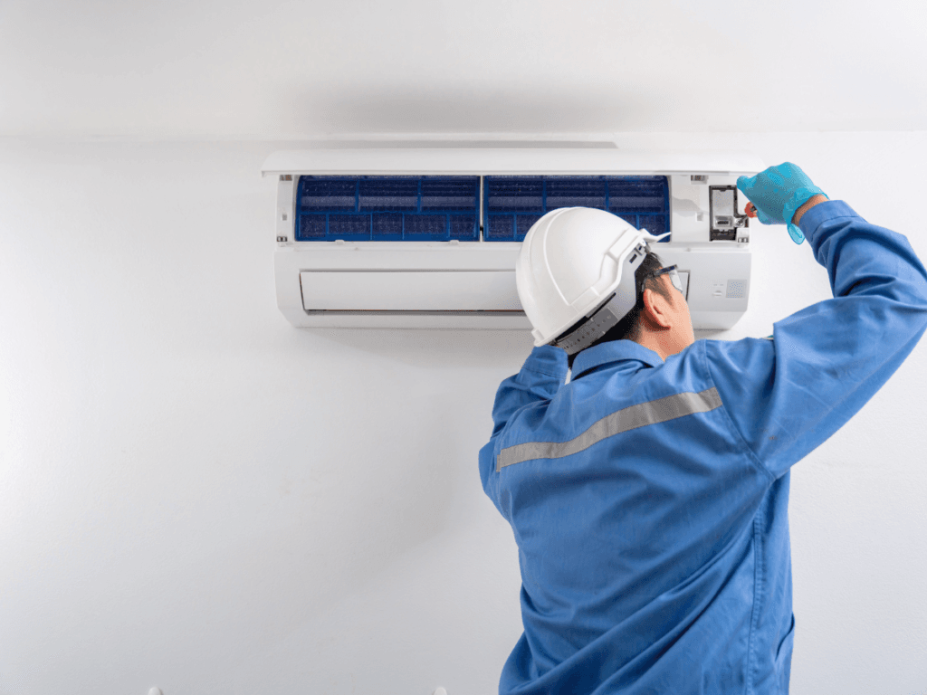 A worker in a blue uniform and white helmet repairs a wall-mounted air conditioner using a tool, addressing an AC not cooling issue while facing the unit.