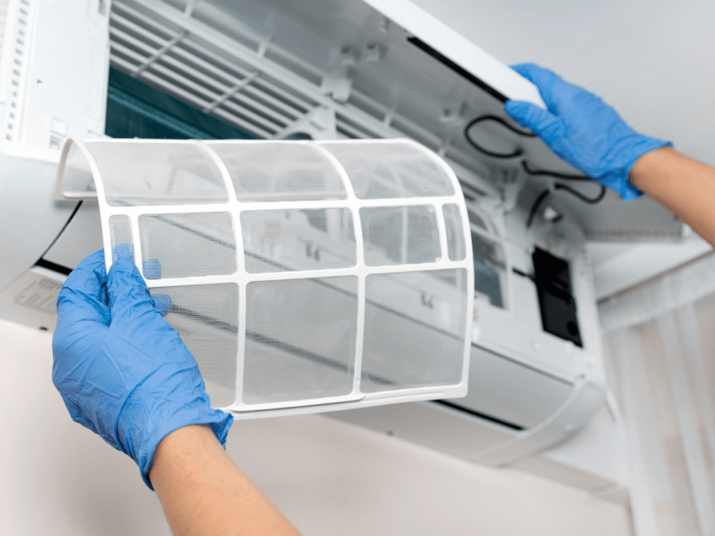 Person wearing blue gloves removes a mesh air filter from a wall-mounted air conditioning unit for cleaning or maintenance, addressing an issue of the AC not cooling properly.