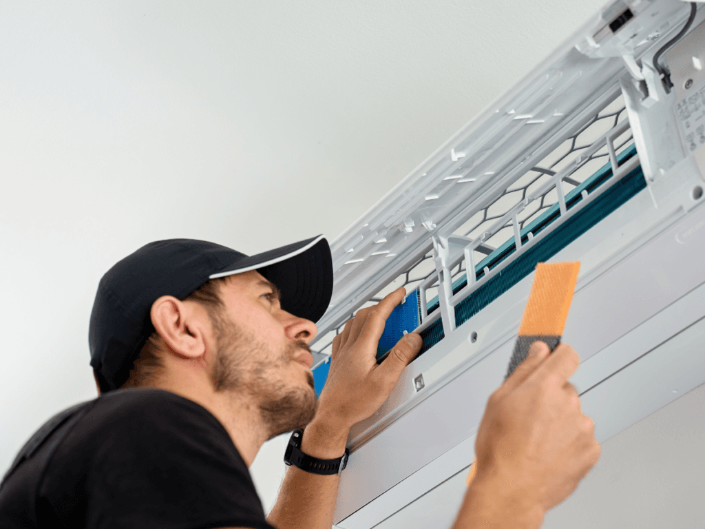 A man in a cap inspects an open wall-mounted air conditioning unit while holding a filter and a cleaning tool, likely troubleshooting why the AC is not cooling.