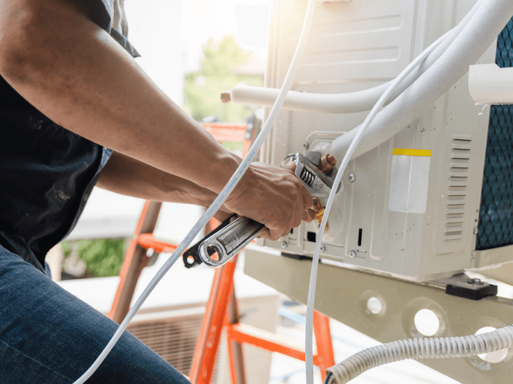 Person using a wrench to adjust or install a clean AC unit outdoors, with hoses and wires visible near the equipment.