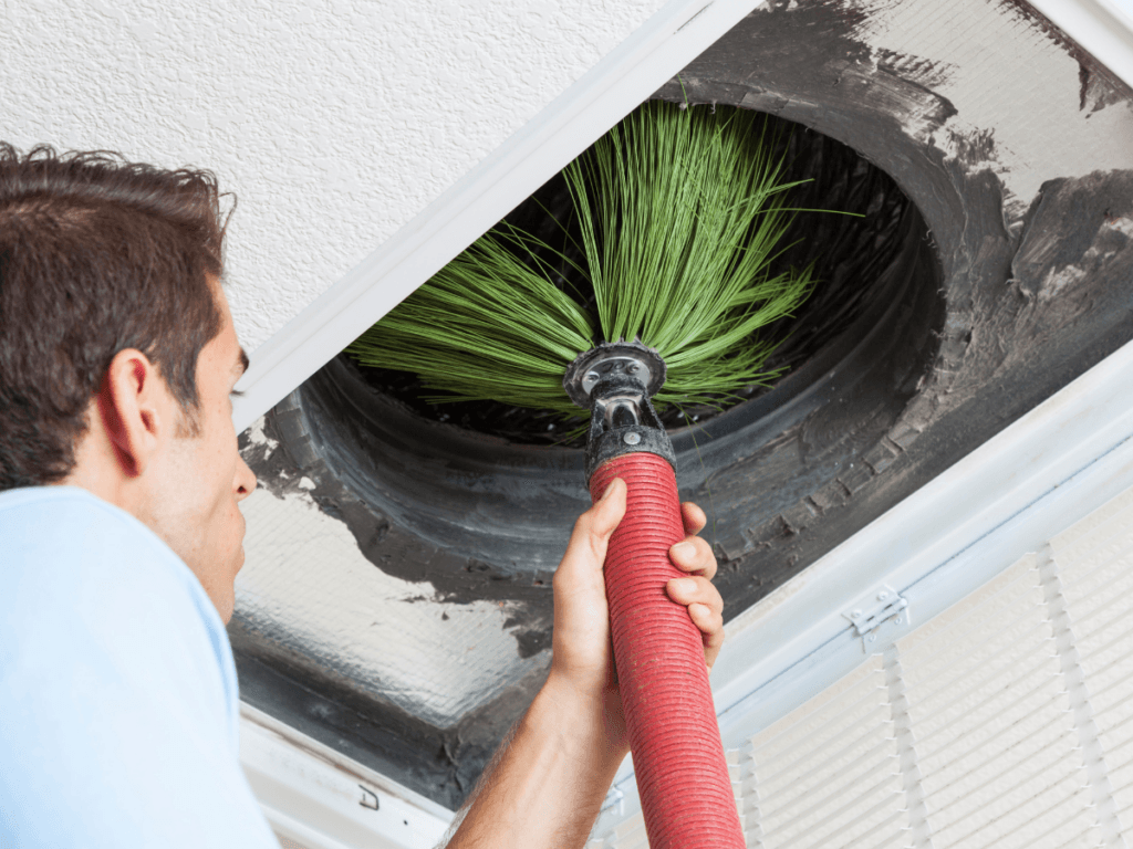 A man uses a brush attached to a hose to clean the inside of an air duct in a ceiling vent, helping to maintain a clean AC unit.