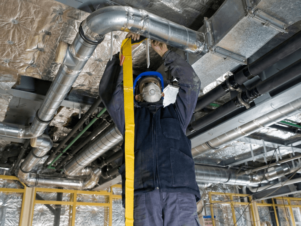 A worker wearing a hard hat and mask adjusts metal ductwork on a ceiling in an industrial building under construction, ensuring proper installation for a clean AC unit.