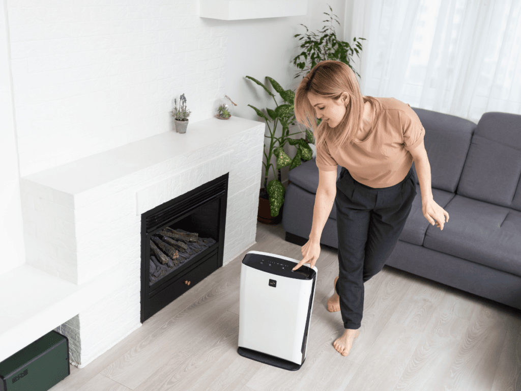 A woman in a living room places her hand on a white air purifier near a fireplace, with plants and a gray sofa in the background, highlighting the comfort and freshness provided by a whole-home dehumidifier.