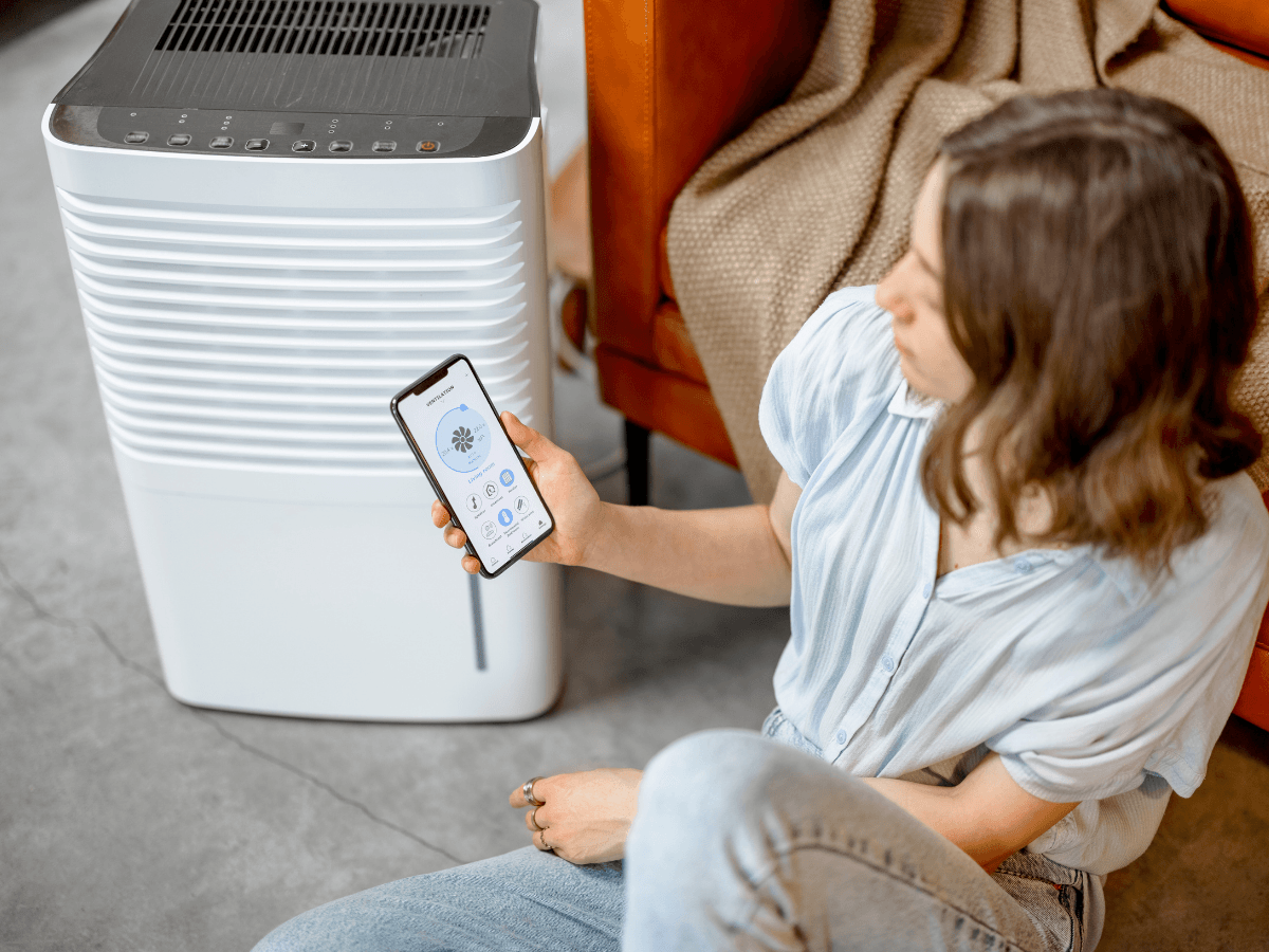 A person sits on the floor, holding a smartphone displaying an app, next to a white whole-home dehumidifier in a modern living space.