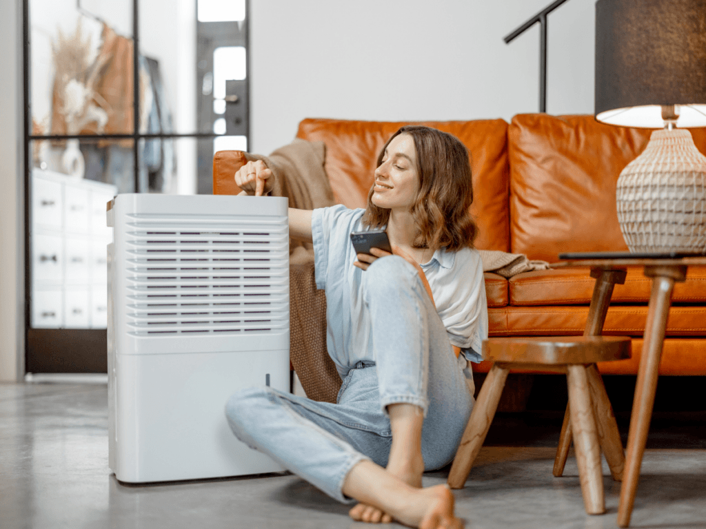 A woman sits on the floor by a brown sofa, adjusting a white whole-home dehumidifier with one hand and holding a remote control in the other.