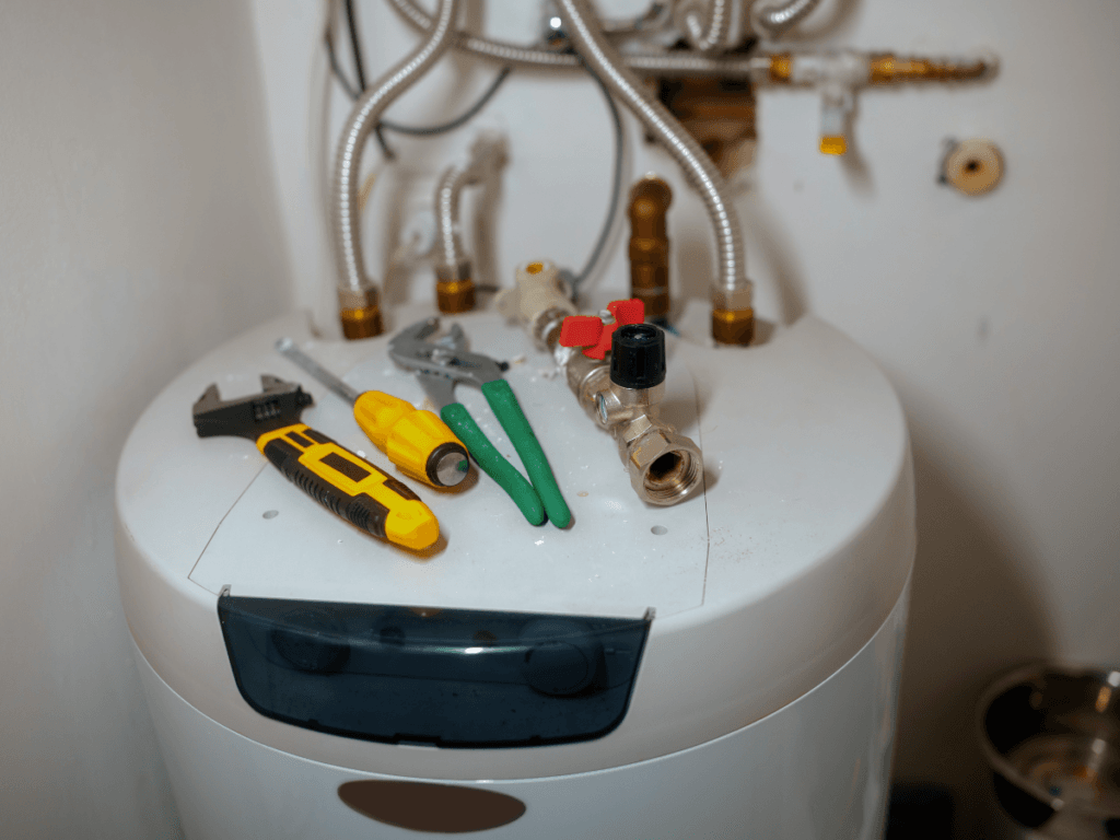A water heater with various hand tools, including pliers, screwdriver, wrench, and utility knife, placed on top—showcasing a traditional repair setup with visible pipes and fittings above.