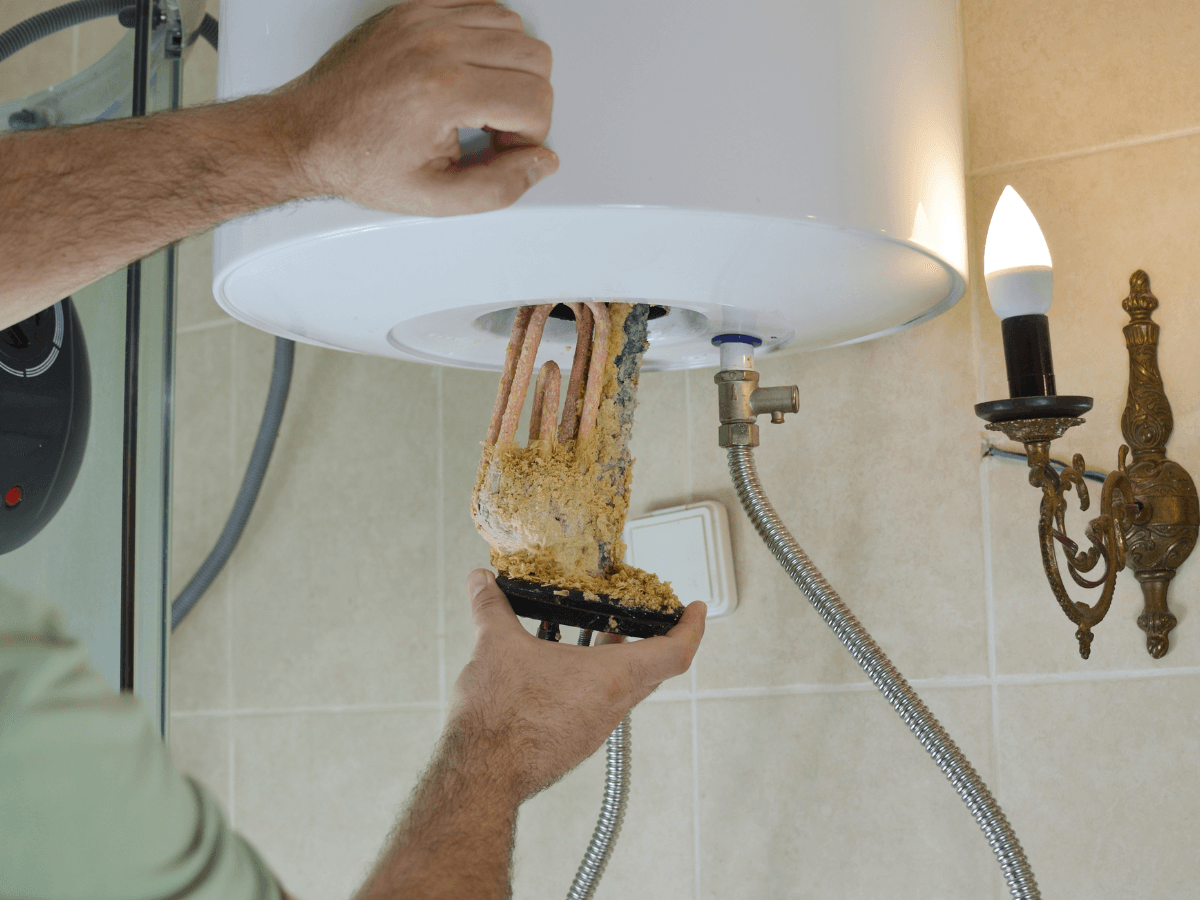 A person performs a traditional repair by removing a heavily scaled heating element from a water heater tank in a bathroom with beige tiles and a wall sconce light.